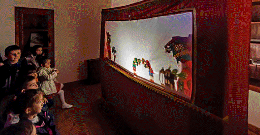 Imagem: Fotografia. Em uma sala escura, muitas crianças sentadas observam um teatro diante de um tecido iluminado. Fim da imagem.