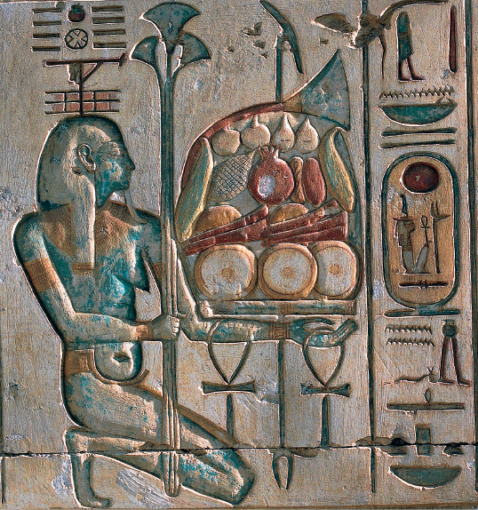 Imagem: Fotografia. Destaque de uma parede com desenhos contornados formando um relevo e com cores desbotadas. Do lado esquerdo, um homem seminu e com adorno na cabeça está ajoelhado e segura uma lança apoiada com a mão direita. Está com o outro braço estendido sobre o qual equilibra uma travessa com muitos alimentos. Deste lado também segura duas cruzes da vida. Ao redor, outros desenhos esparsos. Fim da imagem.