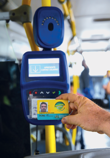 Imagem: Fotografia. No interior de um ônibus, destaque da mão de uma pessoa que aproxima um cartão de plástico de um aparelho eletrônico. Fim da imagem.
