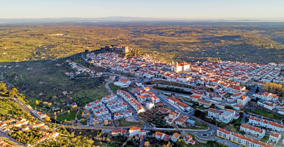 Imagem: Fotografia. Vista aérea de uma pequena cidade rodeada por área verde cultivada e com casas esparsas próximas da cidade. Há uma igreja destacada ao centro e não há construções elevadas.  Fim da imagem.