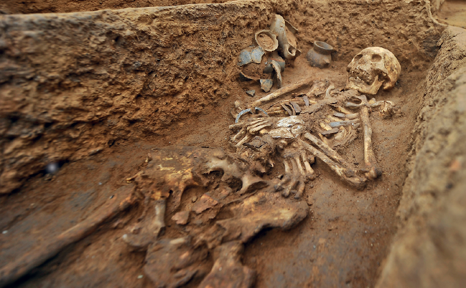Imagem: Fotografia. Um esqueleto humano em uma vala de terra. Fim da imagem.