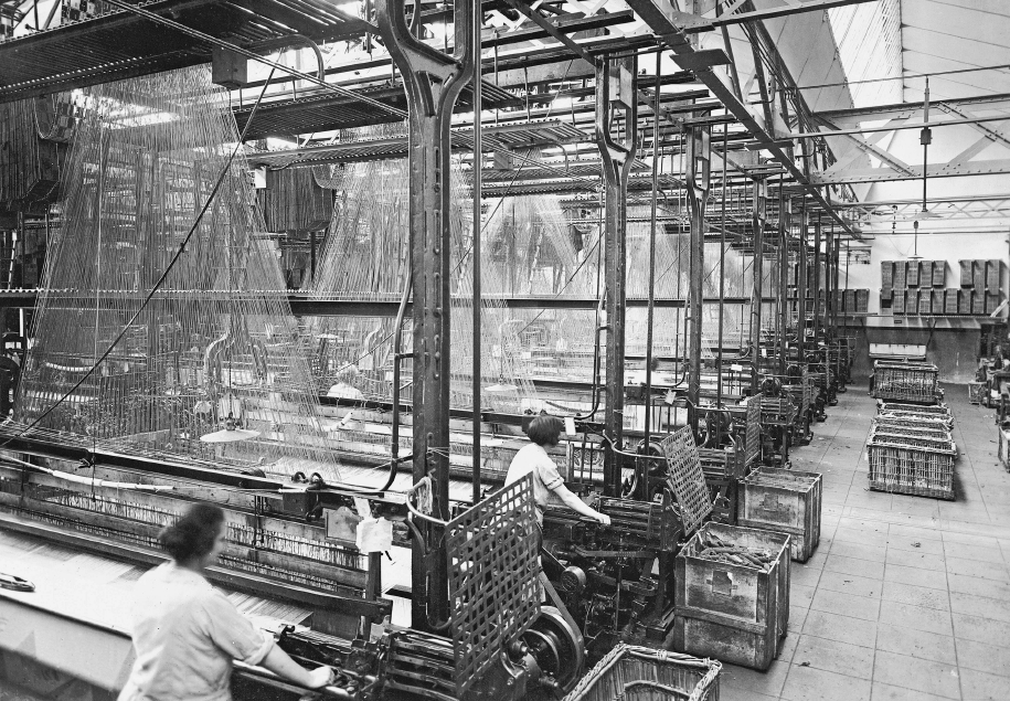 Imagem: Fotografia em preto e branco. Interior de um grande galpão onde mulheres trabalham em grandes máquinas de tear. As estruturas vão do chão ao teto e as linhas são presas e esticadas no vão. Do lado direito, um corredor com caixas.  Fim da imagem.