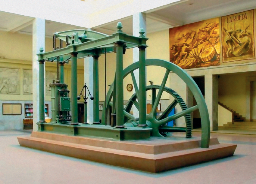 Imagem: Fotografia. Interior de uma sala ampla com pé direito alto e quadros na parede onde está uma grande máquina ao centro. Ela apresenta três pares de barras laterais com uma estrutura pendular ligada a fios no vão entre as barras. Do lado direito, há duas grandes roldanas. Fim da imagem.