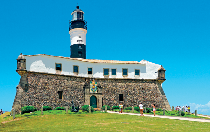 Imagem: Fotografia. Plano aberto que apresenta a fachada de uma robusta parede com porta e janelas com destaque para um farol. À frente do espaço, há algumas pessoas.  Fim da imagem.
