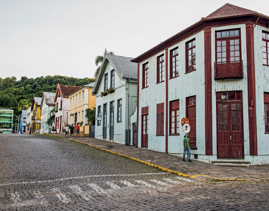 Imagem: Fotografia. Destaque do cruzamento de duas ruas de uma cidade. A via é feita de pedras, há sinalização da faixa de pedestre no chão e construções assobradadas com muitas janelas e telhados em formato de “V” invertido. Fim da imagem.