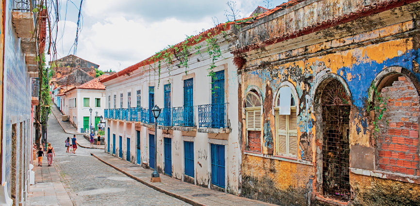 Imagem: Fotografia. Destaque de uma via estreita feita de pedras e calçadas também estreitas. Há construções compridas assobradadas com sacada e portas de acesso direto à rua. Ao fundo, pessoas caminham pelo espaço.  Fim da imagem.