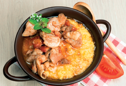 Imagem: Fotografia. Uma panela que apresenta um prato de aspecto úmido com pedaços de carne e pasta amarela. Ao lado, uma colher de madeira e um pedaço de tomate cortado. Fim da imagem.