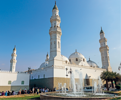Imagem: Fotografia. Fachada de uma construção robusta com quatro compridas torres com detalhes em relevo nas superfícies e estrutura com teto abobadado ao centro. Do lado de fora, há pessoas aglomeradas e uma fonte de água circular.  Fim da imagem.
