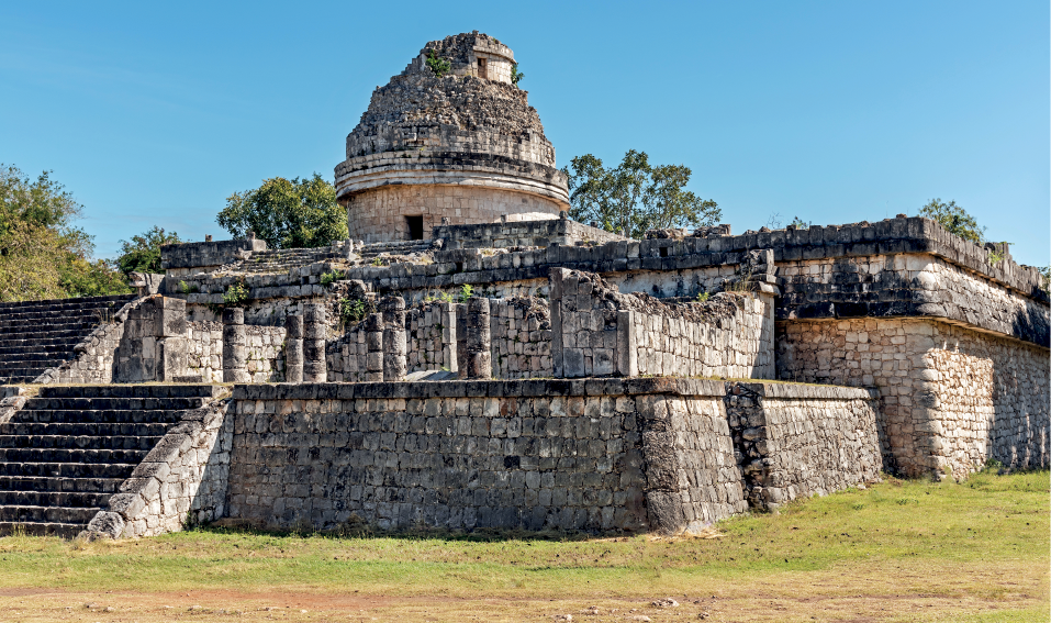 Imagem: Fotografia. Destaque de uma construção em ruínas rodeada por área verde. Apresenta paredes robustas de pedras, tem dois lances de escadas, sendo que no primeiro piso há paredes e colunas destruídas. No piso superior, há uma construção circular com teto abobadado também arruinado.  Fim da imagem.