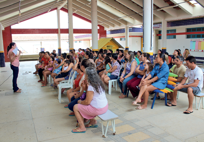 Imagem: Fotografia. Destaque do pátio de uma escola onde homens e mulheres adultos e crianças estão sentados e observam uma mulher falar ao microfone à frente de todos.  Fim da imagem.