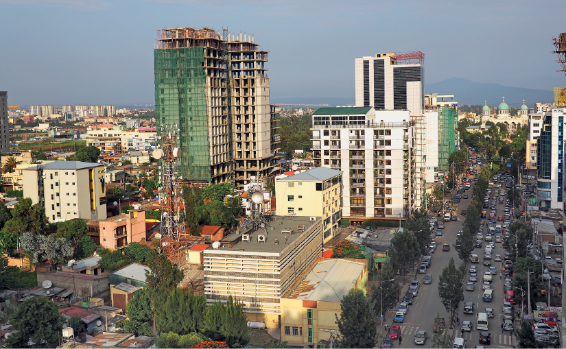 Imagem: Fotografia. Paisagem urbana com uma extensa avenida com muitos carros e muitos prédios ao redor, alguns em construção.  Fim da imagem.