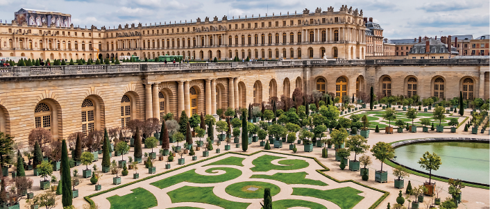 Imagem: Fotografia. Destaque do interior de um espaço que apresenta paisagismo no amplo jardim que é contornado por uma construção extensa, com muitos andares, com janelas arqueadas e fachada com tijolos aparentes.   Fim da imagem.