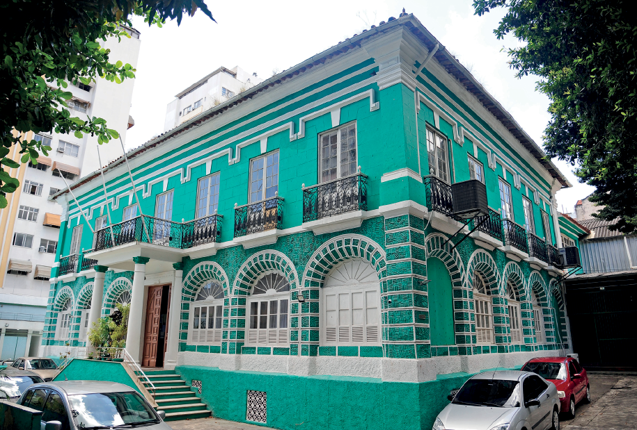 Imagem: Fotografia. Fachada de prédio assobrado com formato retangular e que apresenta muitas janelas arqueadas na parte inferior e janelas retangulares com sacada na parte superior. Na entrada, há uma pequena escada dupla e colunas na área da porta. Ao redor, carros estacionados.  Fim da imagem.