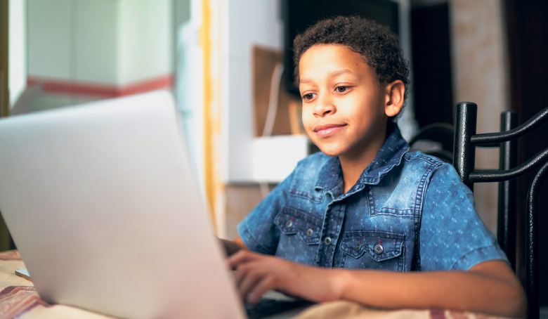 Imagem: Fotografia. Um menino de camisa de manga curta está senado à mesa diante de um notebook aberto.  Fim da imagem.