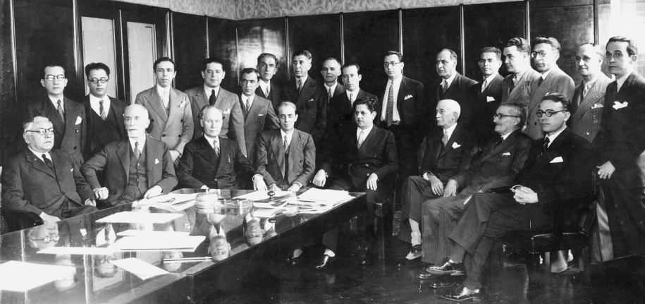 Imagem: Fotografia em preto e branco. Um grupo de 24 homens de terno no interior de uma sala posa para foto. Uma parte está sentada e outros de pé enfileiras na parte de trás. Do lado esquerdo está uma mesa comprida cobre a qual há papéis. Fim da imagem.
