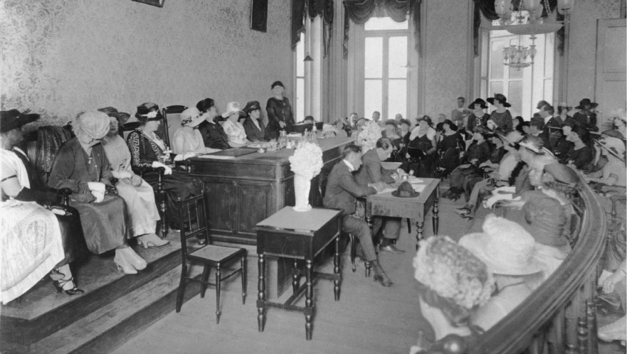 Imagem: Fotografia em preto e branco. Homens e mulheres reunidos em uma ampla sala, a maioria está sentada. Elas ocupam a maior parte da sala. Ao centro, um grupo de homens manipula papéis. As mulheres usam vestidos longos, chapéus e luvas. Os homens usam terno.  Fim da imagem.