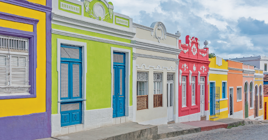 Imagem: Fotografia. Destaque de uma via com calçadas estreitas e com construções térreas que apresentam fachadas coloridas com portas e janelas de madeiro com acesso direto à rua.  Fim da imagem.