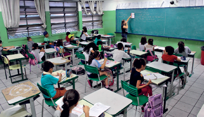 Imagem: Fotografia. Interior de uma sala de aula onde estão alunos crianças sentados diante de carteiras e enfileirados à frente da lousa, onde está a professora.  Fim da imagem.