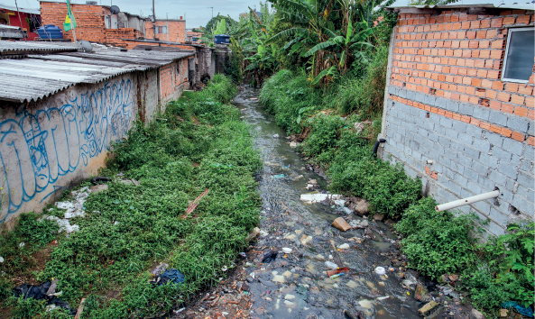 Imagem: Fotografia. Destaque de um córrego com dejetos margeado por arbustos e pequenas casas com paredes sem reboco e caixa d’água no telhado.  Fim da imagem.