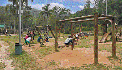 Imagem: Fotografia. Área aberta com chão de terra e grama onde brincam crianças em um parquinho com estrutura de madeira. Ao fundo, vegetação. Fim da imagem.