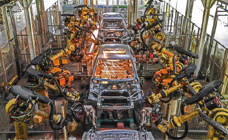 Imagem: Fotografia. Lataria de carros enfileirados sendo soldados por grandes maquinários lado a lado.  Fim da imagem.