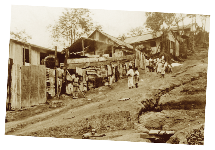Imagem: Fotografia sépia. Destaque de uma ladeira com chão de terra por onde caminham algumas pessoas. No canto direito, uma vala comprida e estreita. Do lado esquerdo, casebres de madeira com muros feitos com pedaços de telhas e ripas também em madeira.  Fim da imagem.